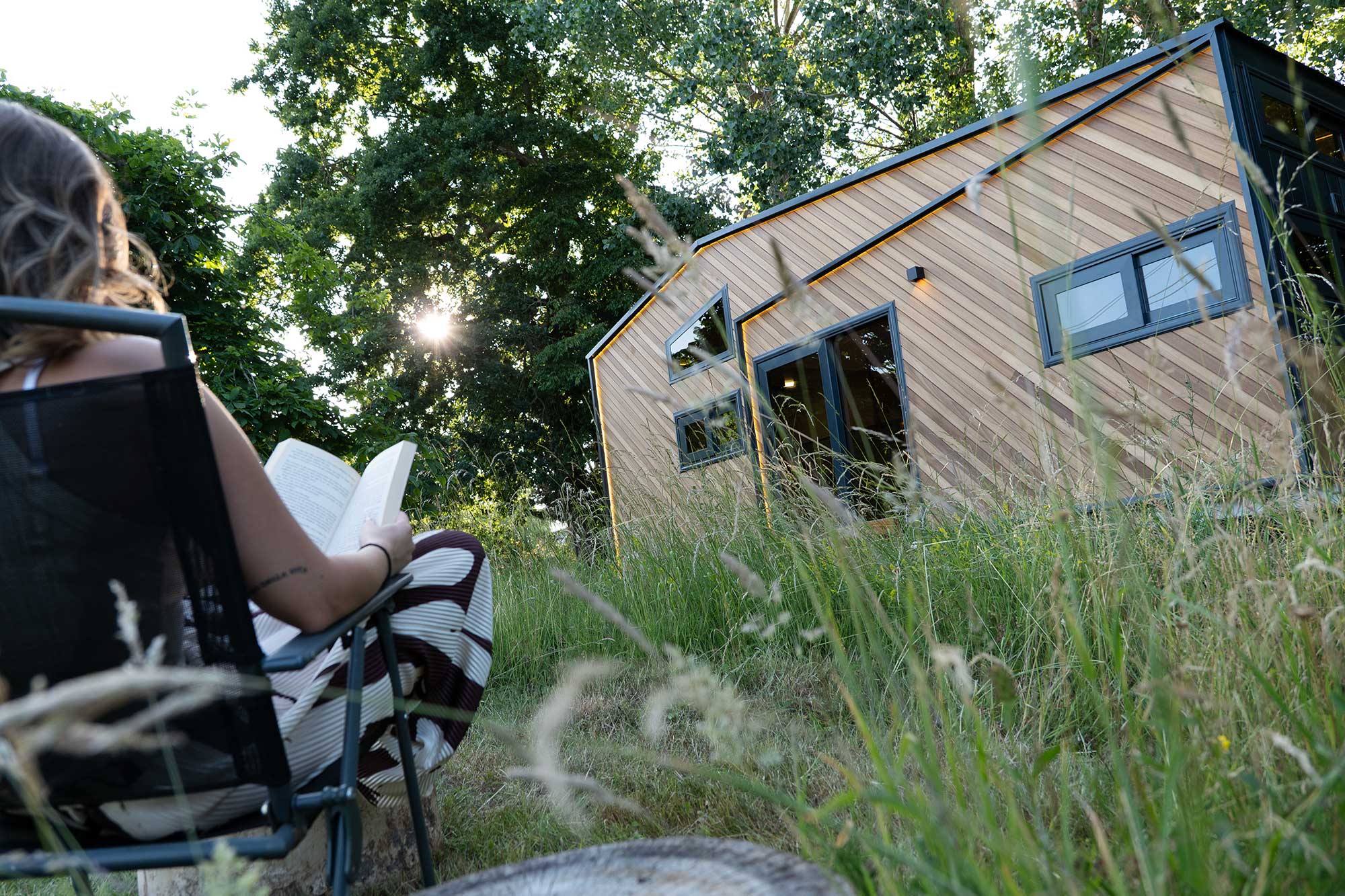 A girl sat reading a book in front of a tiny house on wheels